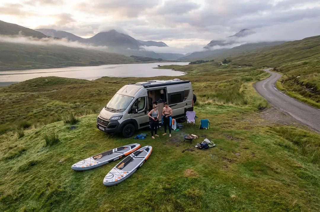 Couple with Loco inflatable SUP boards and campervan parked beside misty Scottish loch on overcast morning