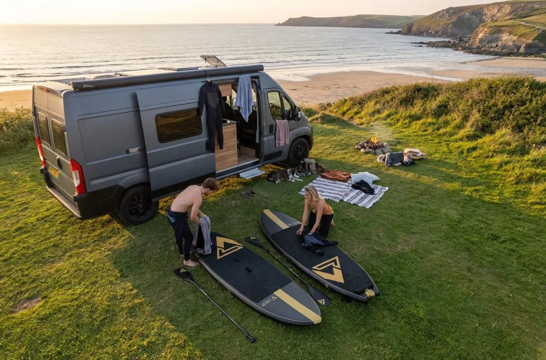 Couple preparing Loco SUP boards beside campervan parked on cliff overlooking secluded UK cove at sunset