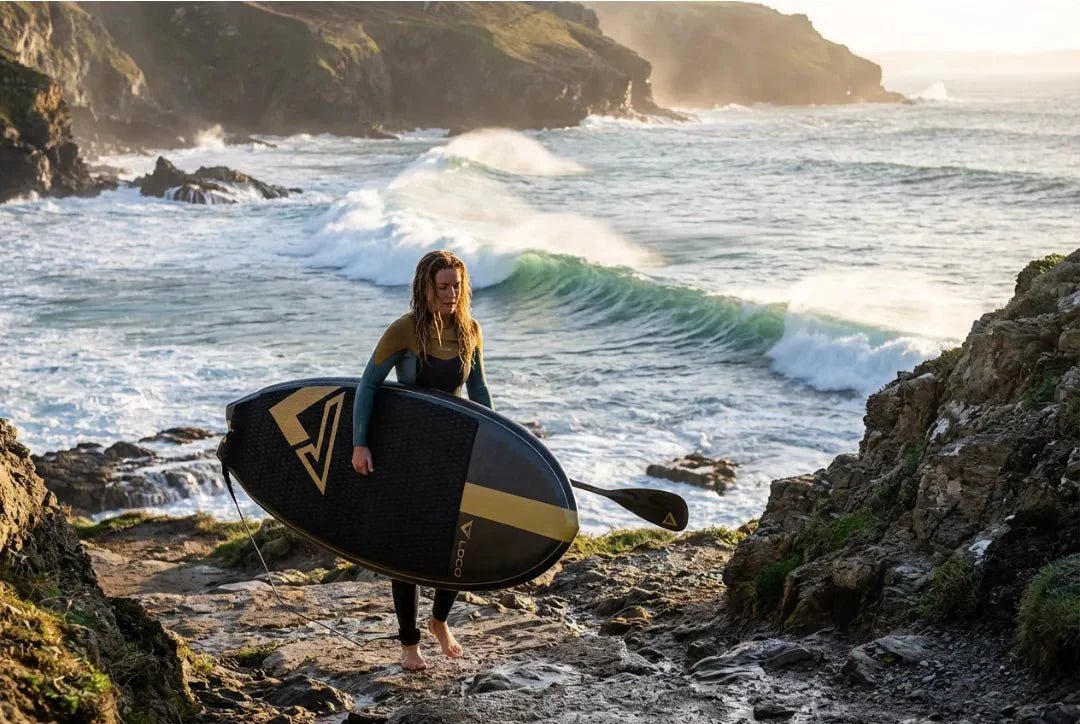 Female SUP surfer in wetsuit carrying Loco hard board down rocky cliff path to UK surf break at golden hour