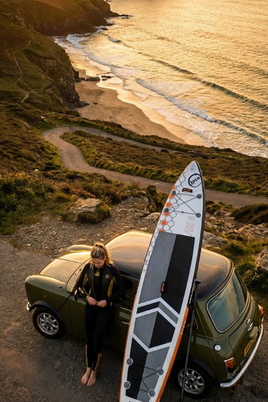 Girl holding a SUP board next to a vintage car on a coastal road at sunset.