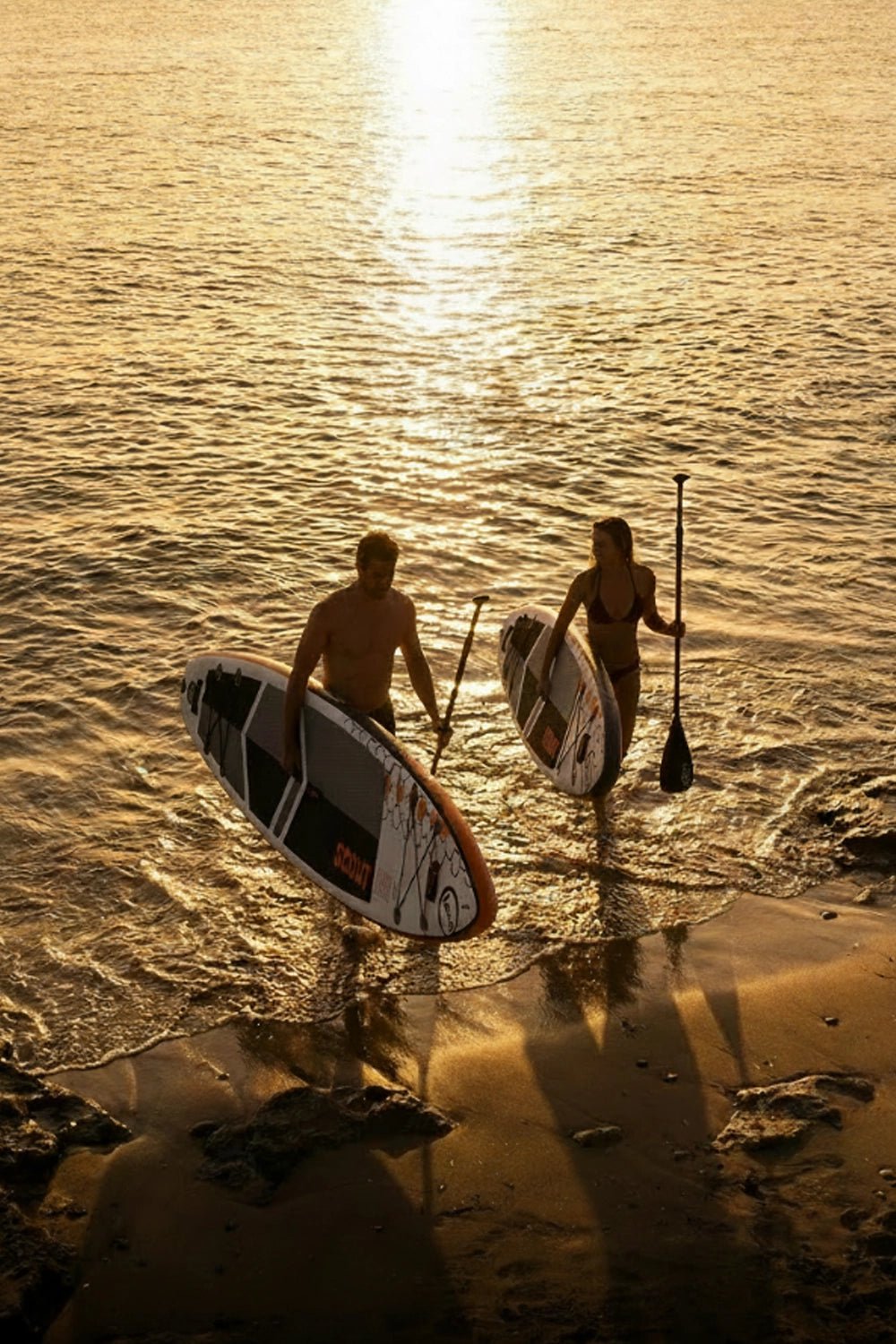 Two people with paddleboards walking out of the water at sunset.
