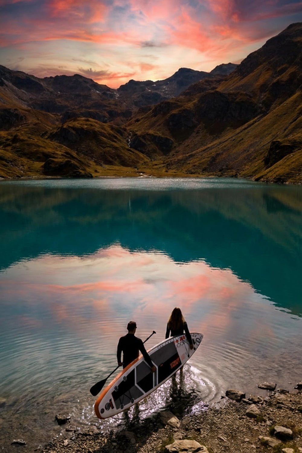 Two people with a paddleboard on a mountain lake at sunset.