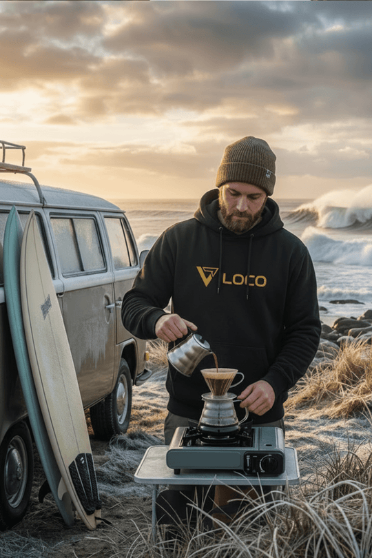 Surfer making coffee with campervan and waves in background on a fresh but sunny day