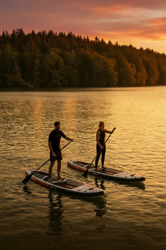 Two people paddleboarding on a lake at sunset with trees in the background.