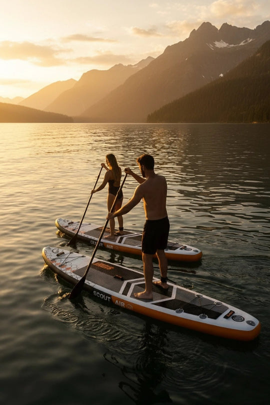 Two people paddleboarding on a lake with mountains in the background during sunset.