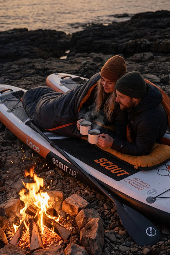 Two people sitting on a Scout Air paddleboard by a campfire on a rocky shore.
