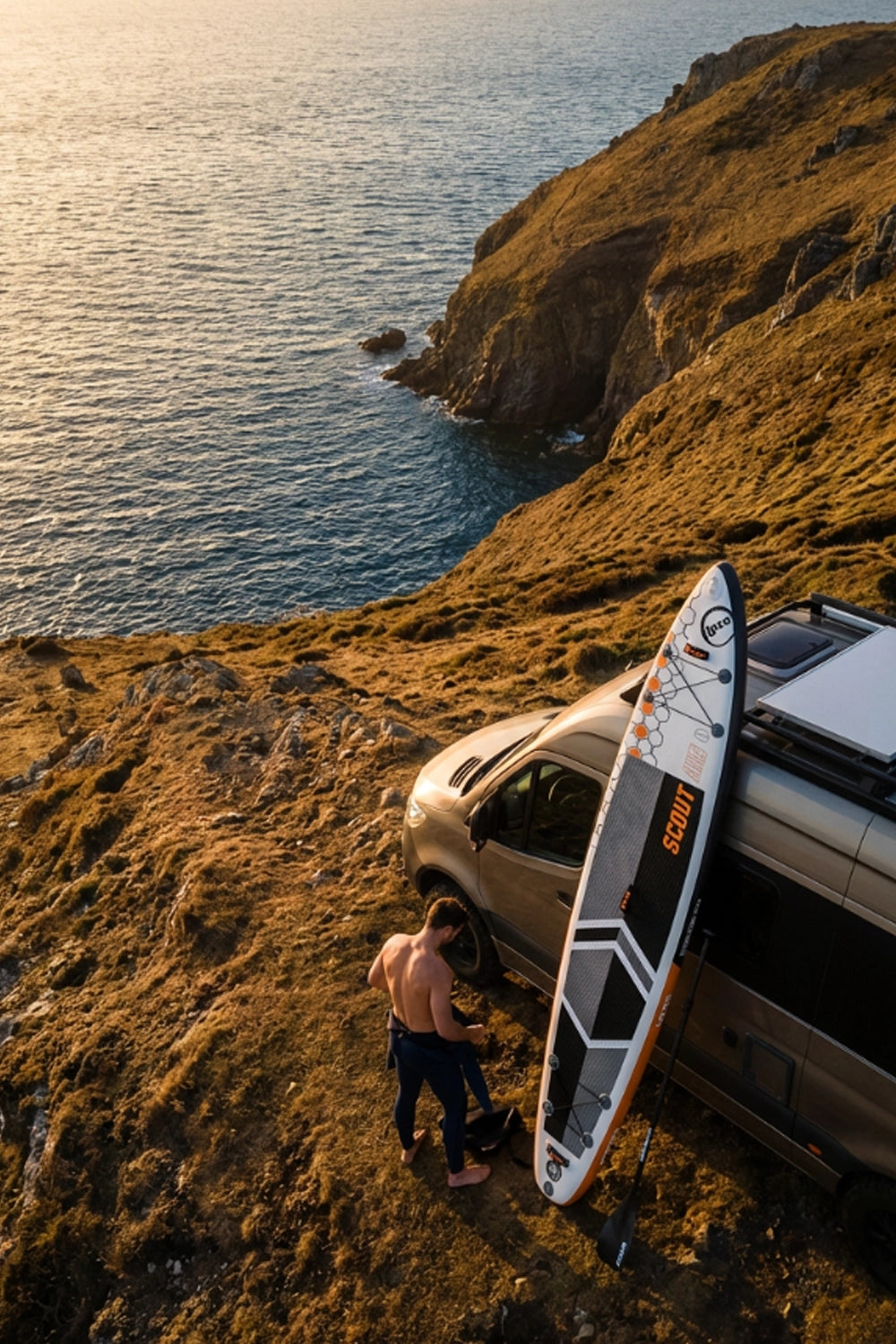 Person with a touring SUP board near a van on a grassy hill overlooking the ocean.