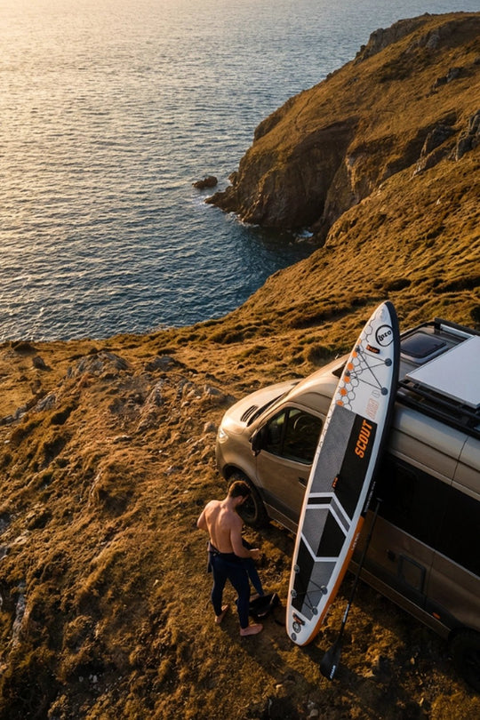 Person with a touring SUP board near a van on a grassy hill overlooking the ocean.