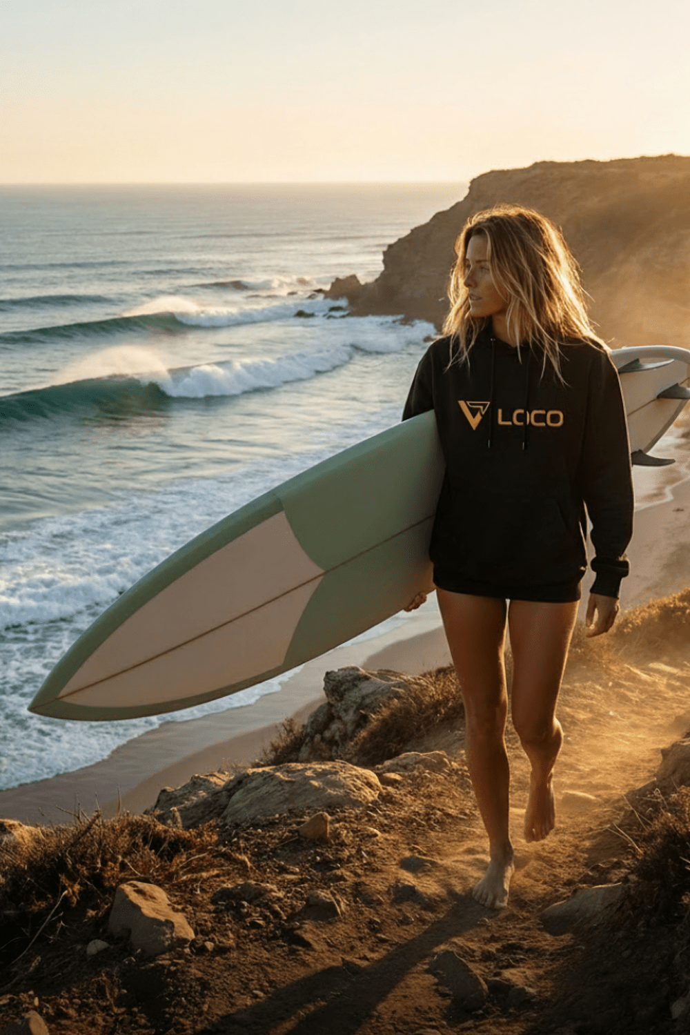 Paddle surfing woman walking on a beach with a surfboard, wearing a black Loco hoodie 