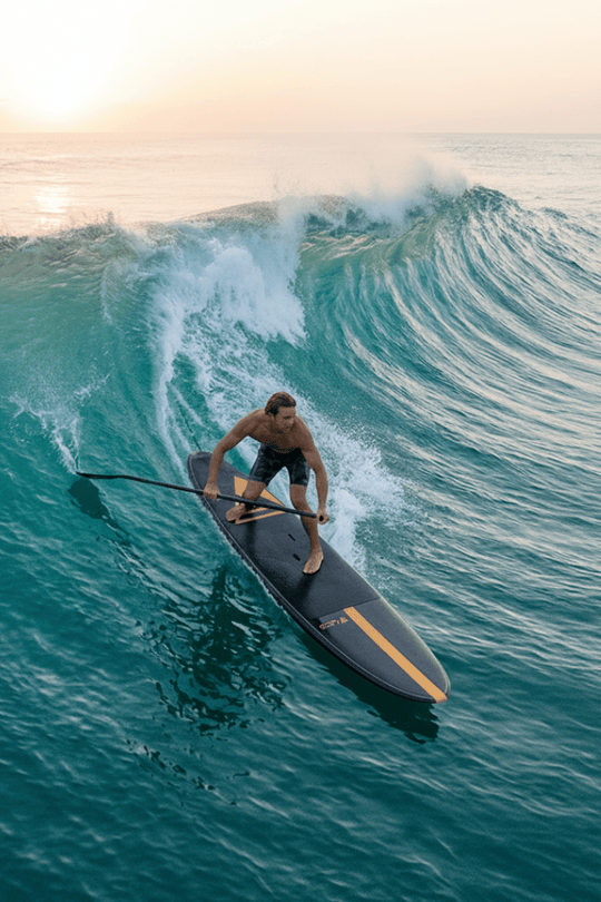 Guy SUP surfing a longboard on a large wave in the ocean at sunset.